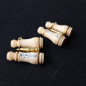Puede incluir: Cuatro gemelos antiguos de color marfil con detalles metálicos dorados. Cada gemelo tiene forma cilíndrica con una base ligeramente ensanchada y un conector metálico dorado. Los gemelos tienen una estética vintage y están sobre una superficie oscura.