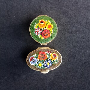 Puede incluir: Dos pequeñas cajas de baratijas de mosaico vintage. Una es redonda, la otra ovalada. Ambas presentan diseños florales en rojo, amarillo, azul y blanco sobre un fondo verde. Las cajas tienen marcos de metal color dorado.