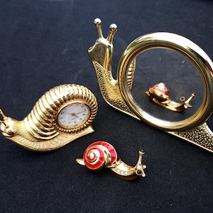 Op de afbeelding: Een verzameling goudkleurige slakkenbeeldjes op een zwarte ondergrond. Eén is een klok, een andere een spiegel en de derde een broche met rode en witte accenten. De klok heeft een witte wijzerplaat met zwarte cijfers.