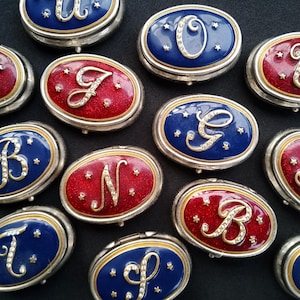 Puede incluir: Una variedad de joyeros ovalados con bordes plateados. Las cajas presentan tapas de esmalte rojo o azul, cada una adornada con una letra brillante y pequeños detalles de estrellas. Las letras están adornadas con pedrería.