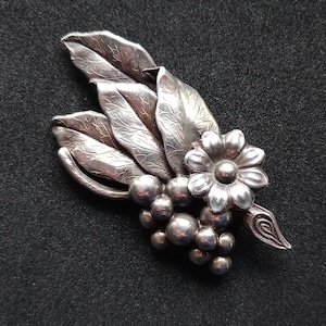 Peut inclure: Broche argentée représentant un motif floral et végétal. La broche comprend un groupe de formes rondes, une fleur et plusieurs feuilles détaillées. La broche est posée sur un fond sombre.