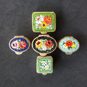 Puede incluir: Cinco pequeñas cajas cuadradas y ovaladas con diseños florales en mosaico. Las cajas son de metal y tienen un borde dorado. Las flores son en tonos de rojo, naranja, amarillo, verde, azul y blanco.