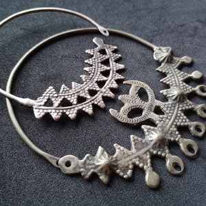 May include: A pair of silver hoop earrings with intricate, geometric designs. The earrings are made of two hoops, one larger than the other, with a decorative element attached to the larger hoop.