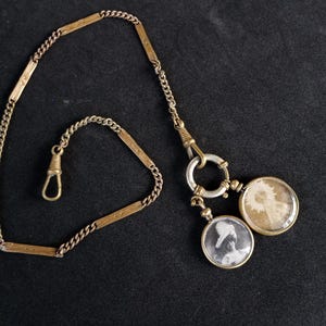 Puede incluir: Cadena de reloj de bolsillo antigua con dos medallones circulares. La cadena es dorada con un cierre de gancho. Un medallón contiene un retrato en blanco y negro, el otro un diseño floral. La cadena del reloj se muestra sobre una superficie oscura.
