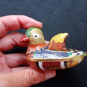 Puede incluir: Una pequeña caja de baratijas de cloisonné con forma de pato que presenta intrincados patrones de esmalte en tonos vibrantes de rojo, azul, verde, amarillo y blanco. La tapa con borde metálico tiene forma de cabeza y ala de pato.