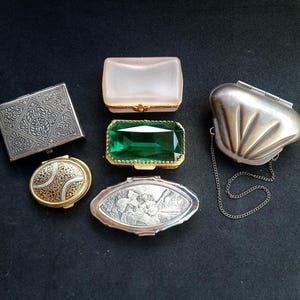 Puede incluir: Una colección de cajas antiguas y ornamentadas de varias formas y tamaños. Incluye una caja cuadrada plateada con patrones detallados, una caja ovalada dorada y plateada, y un bolso plateado en forma de concha con una cadena.
