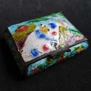Puede incluir: Una pequeña caja rectangular con bisagras y un diseño de esmalte colorido. La caja está decorada con flores, pájaros y una escena de paisaje. La caja está hecha de metal y tiene una bisagra de latón.