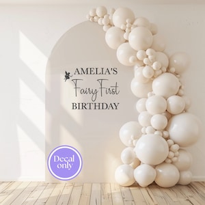 Könnte beinhalten: Ein großer, bogenförmiger Aufkleber mit dem Text "AMELIA'S Fairy First BIRTHDAY" in schwarzer Schrift, begleitet von einer Feenillustration. Eine Ansammlung cremefarbener Ballons schmückt die rechte Seite. Der Aufkleber ist mit "Decal only" in einem lila Kreis versehen.