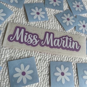Könnte beinhalten: Ein rechteckiger Aufkleber mit dem Text Miss Martin in violetter Schreibschrift und weißer Umrandung, umgeben von mehreren kleineren quadratischen Aufklebern, die jeweils eine weiße Gänseblümchenblüte mit violettem Zentrum auf hellblauem Grund zeigen.