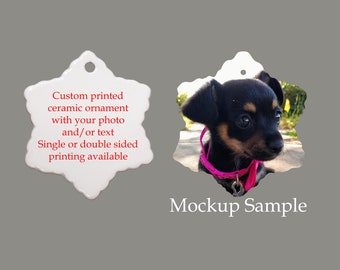 Puede incluir: Un adorno de cerámica blanco en forma de copo de nieve con una imagen personalizada impresa de un perro pequeño negro y marrón con un collar rosa. El adorno está en blanco en el otro lado y tiene un agujero para colgar. El texto "Custom printed ceramic ornament with your photo and/or text Single or double sided printing available" está impreso en el lado en blanco. El texto "Mockup Sample" está impreso debajo del adorno.