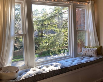 Könnte beinhalten: Ein Fensterplatz mit einem grauen Kissen und weißem eingebautem Stauraum. Das Fenster hat weiße Vorhänge und eine Lichterkette, die darüber hängt. Das Fenster blickt auf einen grünen Baum und eine Straße.