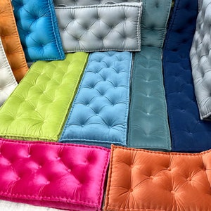 Op de afbeelding: Assortiment van gestoffeerde vloer kussens in verschillende kleuren, waaronder groen, blauw, roze, oranje en grijs. De kussens zijn rechthoekig en hebben een knoopsteek ontwerp.