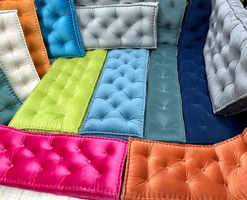 Op de afbeelding: Assortiment van gestoffeerde vloer kussens in verschillende kleuren, waaronder groen, blauw, roze, oranje en grijs. De kussens zijn rechthoekig en hebben een knoopsteek ontwerp.