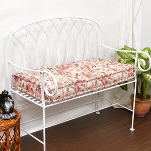 May include: A white metal bench featuring a curved backrest with a lattice design and scrolled armrests. The bench includes a rectangular seat cushion with a beige and pink floral pattern, providing a soft surface for the metal frame structure.