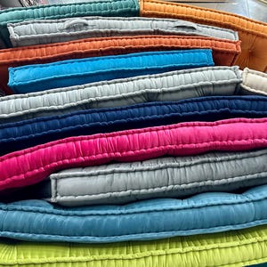 May include: A stack of rectangular floor cushions in various colors including teal, orange, light gray, bright blue, dark blue, hot pink, and lime green. Each cushion features a tufted design with visible stitched edges and a soft fabric texture.
