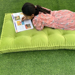 Op de afbeelding: Een groene, gestoffeerde vloercushion met een persoon die erop ligt en een boek leest. De cushion heeft een dikke, gevulde rand.