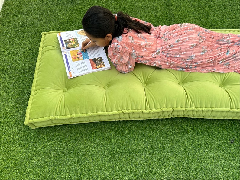 Op de afbeelding: Een groene, gestoffeerde vloercushion met een persoon die erop ligt en een boek leest. De cushion heeft een dikke, gevulde rand.
