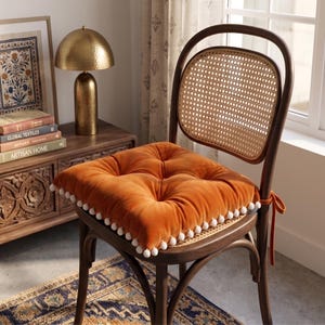 May include: A square, tufted orange velvet seat cushion featuring white pom-pom trim along the edges and fabric ties on the side. The cushion is placed on a dark wood chair with a curved backrest and a woven cane insert in the center.