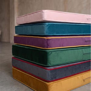 Puede incluir: Una pila de seis cojines de suelo rectangulares hechos de tela de terciopelo. Los cojines son de color rosa claro, azul cerceta, morado con ribete amarillo, verde oscuro, gris oscuro con ribete rojo y amarillo mostaza, cada uno con cremallera.