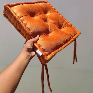 Op de afbeelding: Een vierkant, getuft oranje fluwelen zitkussen met een zachte, pluche textuur en vier opvallende knoop-getufte inkepingen. Het kussen is voorzien van bijpassende oranje stoffen linten op de hoeken en een wit label met de tekst QUILTELLA.
