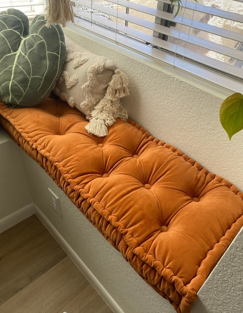 Op de afbeelding: Een groot, oranje, gestoffeerd kussen met een dikke, fluweelachtige stof. Het kussen ligt op een vensterbank en heeft een decoratief, gefranst kussen ervoor.