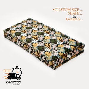 May include: A large, rectangular floor cushion with a geometric pattern in shades of green, yellow, pink, and brown. The cushion is made of fabric and has a tufted design. The text "CUSTOM SIZE... SHAPE... & FABRICS..." is visible in the upper right corner of the image.