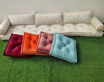 May include: A long, cream-colored tufted floor sofa with three rectangular back cushions and two cylindrical side bolsters. In front, four square tufted floor cushions are arranged in a row, colored in deep magenta, orange, dusty rose, and light blue.