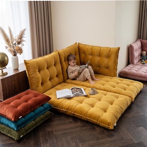 May include: A modular mustard yellow floor sofa with a tufted velvet texture, featuring a corner backrest configuration. A stack of three additional floor cushions in rust, teal, and olive green sits nearby, alongside an open book and a small cup.
