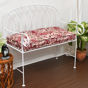 May include: A white metal garden bench with a floral cushion. The cushion has a pattern of red, pink, and cream flowers. The bench features a curved back and scrollwork armrests. It sits on a dark wood floor. The bench is approximately 120 cm long.