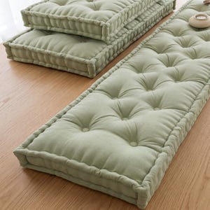 May include: A set of rectangular sage green floor cushions featuring a tufted design with multiple fabric-covered buttons across the surface. The cushions have thick, piped edges and a soft, uniform texture, providing a structured appearance.