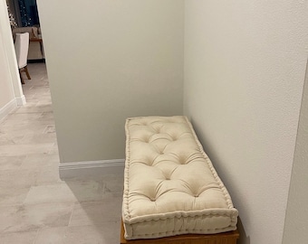 Pode incluir: Um banco de armazenamento retangular de madeira coberto com uma almofada espessa de cor creme com botões. Acima do banco, uma placa de parede retangular apresenta uma moldura de madeira escura, fundo branco, a palavra "home" e coordenadas.