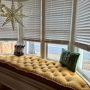 May include: A mustard yellow tufted window seat cushion with a square white pillow featuring a reindeer pattern and a rectangular green pillow with embroidered Christmas tree designs, positioned beneath a hanging white star light fixture.