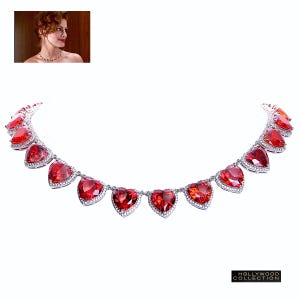 May include: A silver necklace featuring a row of red heart-shaped stones. Each stone is surrounded by smaller clear stones.