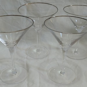 Op de afbeelding: Vier heldere martiniglazen met zilveren randen. De glazen hebben een klassieke driehoekige komvorm en een lange, slanke steel. De basis van elk glas is rond. De glazen zijn gerangschikt op een wit oppervlak.