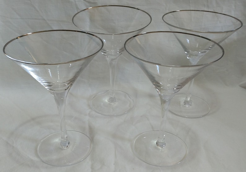 Op de afbeelding: Vier heldere martiniglazen met zilveren randen. De glazen hebben een klassieke driehoekige komvorm en een lange, slanke steel. De basis van elk glas is rond. De glazen zijn gerangschikt op een wit oppervlak.