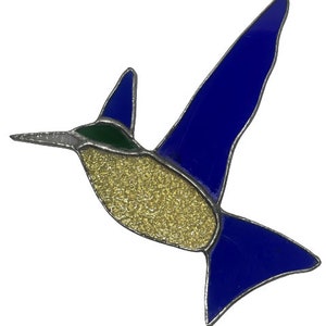 May include: A stained glass hummingbird in flight. The bird features a gold textured body, blue wings and tail, a green head, and a silver beak. The glass is outlined with a silver metal. The hummingbird is a decorative item.