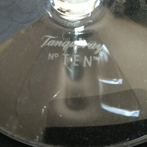 Op de afbeelding: Close-up van de voet van een helder glas met de witte tekst "Tanqueray No TEN" gegraveerd. Het glas is waarschijnlijk een cocktailglas, dat het logo van het merk toont.