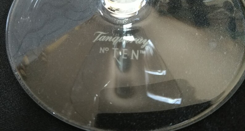Op de afbeelding: Close-up van de voet van een helder glas met de witte tekst "Tanqueray No TEN" gegraveerd. Het glas is waarschijnlijk een cocktailglas, dat het logo van het merk toont.