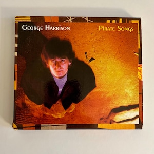 May include: A CD case featuring George Harrison's album "Pirate Songs." The cover has a warm, orange and brown background with a portrait of Harrison. The album title and artist's name are printed in white.