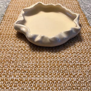 May include: A white ceramic dish with a ruffled edge. The dish is sitting on a brown and white woven placemat.
