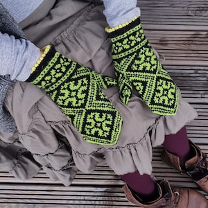 Op de afbeelding: Een paar gebreide wanten met een zwart en neon-groen geometrisch patroon met een herhalend ruitmotief. De wanten hebben een geribbelde boord met een gele rand en zijn gemaakt van dik, getextureerd garen in een traditionele stijl.