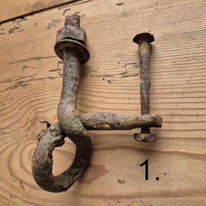 May include: Two pieces of weathered, rusty metal hardware. One has a bolt, nut, and washer, with a curved hook. The other is a bolt with a flat head and a connecting piece. The number "1." is visible.