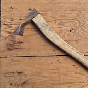 Op de afbeelding: Een vintage dissel met een verweerde, roestige metalen kop met een gebogen blad, bevestigd aan een lange, lichtgekleurde houten steel. Het gereedschap vertoont tekenen van ouderdom en gebruik, met een zichtbare houtnerf en een donkere, geoxideerde patina.