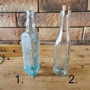 Op de afbeelding: Twee heldere glazen flessen met verschillende vormen. Eén is een vierkante fles met een lichtblauwe tint, en de andere is een lange, slanke fles met een kurk. De flessen staan op een houten oppervlak.