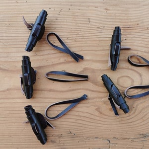 Könnte beinhalten: Ein Set aus vier schwarzen Knebelverschlüssen, die jeweils aus einem zylindrischen schwarzen Bauteil mit einem mittigen Schlitz und einer befestigten schwarzen Lederschlaufe bestehen. Die Knebel sind verstreut angeordnet und zeigen die glatte Oberfläche.
