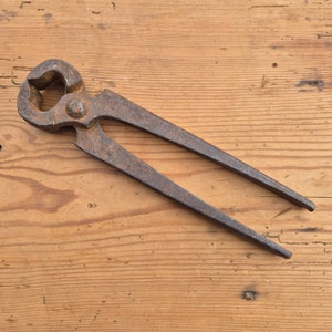 May include: A well-worn farrier's nail puller, showing signs of age, rests on a wooden surface. The tool features a curved head, a central pivot, and long, tapered handles. The metal has a rusty appearance.