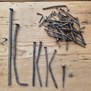 May include: A selection of old, rusty nails in different sizes and forms, including long spikes and smaller nails with flat heads. The nails are displayed on a light brown wooden surface, highlighting their aged look.