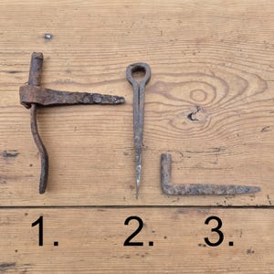 Peut inclure: Trois outils anciens en fer avec une patine rouillée marquée. L'objet 1 est une penture en T, l'objet 2 est une longue pointe métallique fine avec une tête en boucle circulaire, et l'objet 3 est une équerre en métal. Chaque pièce montre de l'usure.
