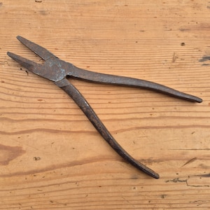 Op de afbeelding: Een vintage metalen tang met een donkere, verweerde en geoxideerde afwerking. Het gereedschap heeft twee lange, taps toelopende bekken en twee gebogen handgrepen die verbonden zijn door een centraal scharnierpunt, met zichtbare gebruikssporen.