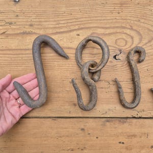 Op de afbeelding: Drie donkere, verweerde, handgesmede ijzeren S-haken met een ruw, getextureerd oppervlak. Eén haak is groot en gebogen, een andere is bevestigd aan een ronde ijzeren ring en de derde is een kleinere, eenvoudige S-vormige haak met ouderdomssporen.